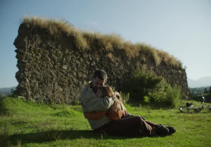 A man holds his daughter while sitting on grass near a rock wall