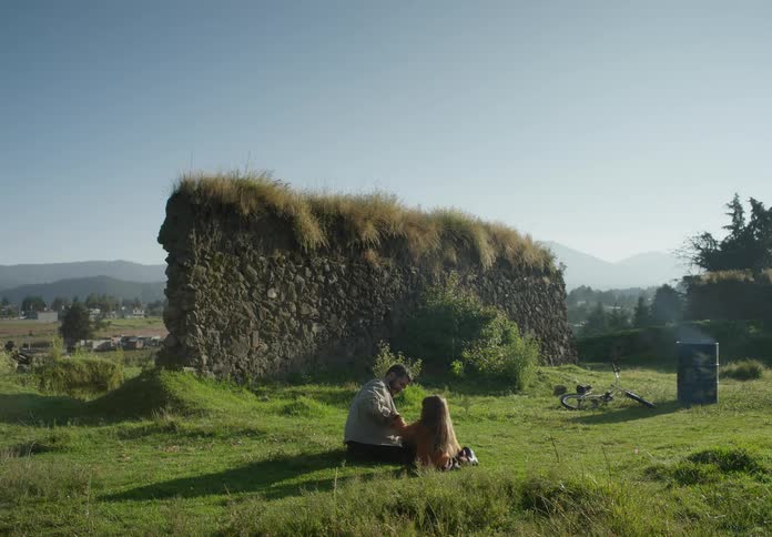 A man is sitting outside in the grass with his young daughter near ruins of a wall and there are mountains in the distance
