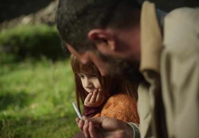 A young girl sits next to her dad outside and he touches her nose with roll of paper