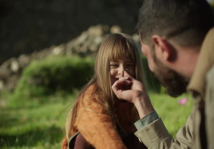 A little girl sits outside in the grass with her dad and they laugh as he puts something small up to her nose