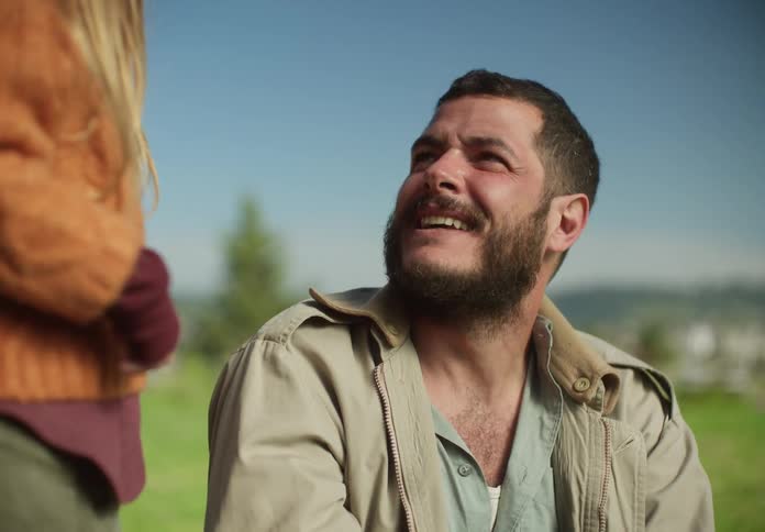 A man is sitting outside in the grass as he looks up and laughs at his young daughter standing next to him and raises his hand as she high fives him then he lights a lighter