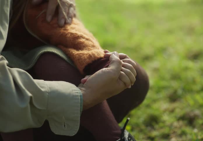 A man sitting on the grass lifts his young daughter in to his lap and they hold hands