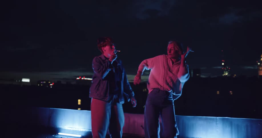 Two women dance as they share cigarettes with city lights in the background