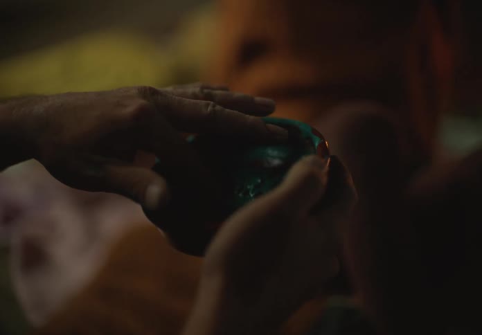 A young girl sits in front of a fireplace as a man rubs his finger in paint and she leans away from him when he goes to put it on her nose