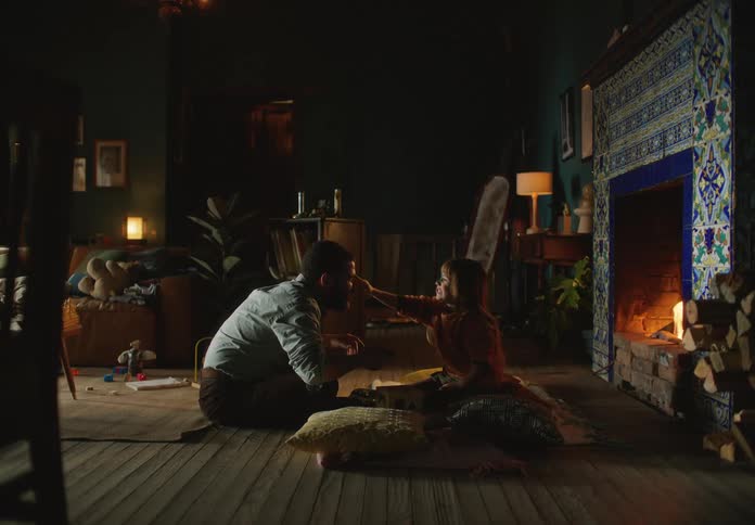 A father and daughter sit at home in front of a burning fireplace and she pretends to feed him