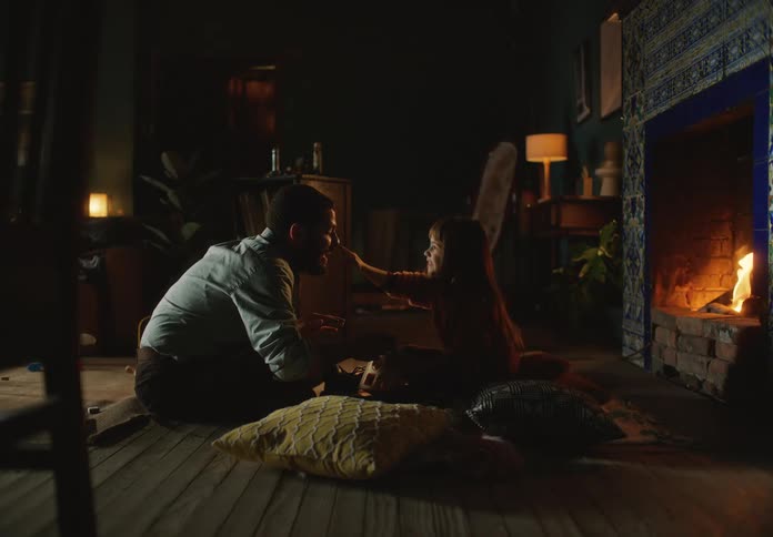 A father and his young daughter sit on the living room floor in front of the fire and the daughter pokes the fathers nose