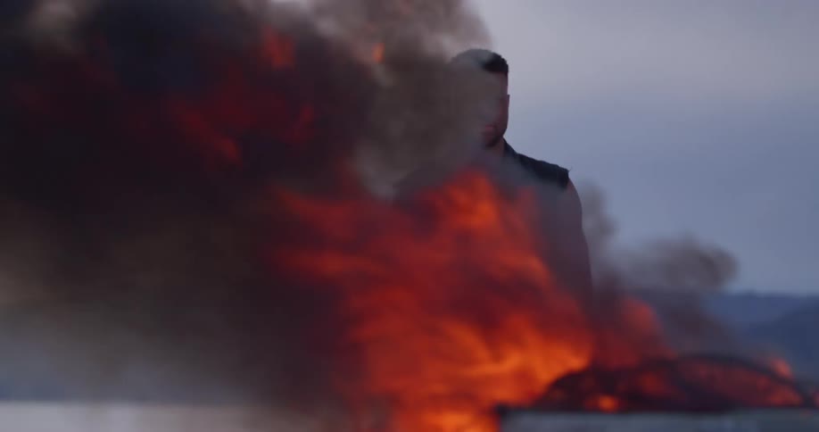 A man is standing outside watching a blazing fire as an automobile burns and black smoke rises from the flames