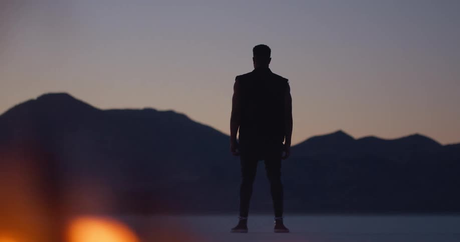 A man standing in the desert faces the distant mountains as a fire burns behind him at sunset