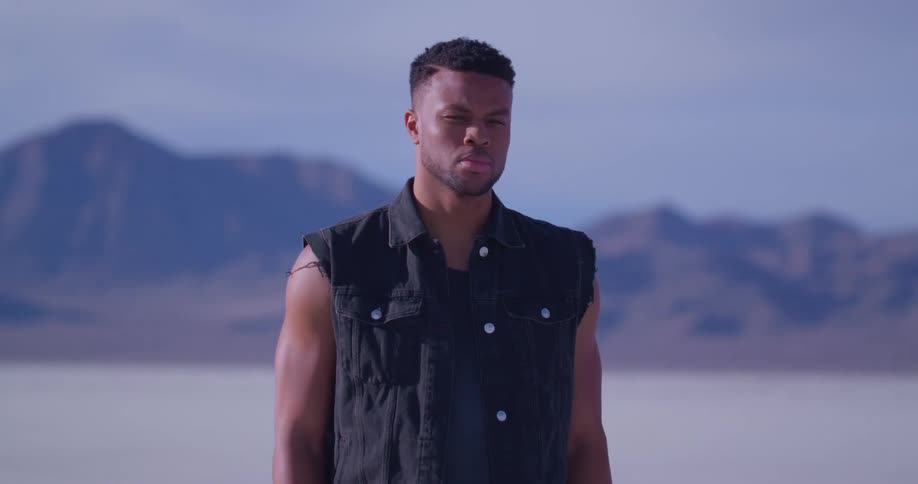 An angry looking man stands in front of a mountain range and looks in to the distance