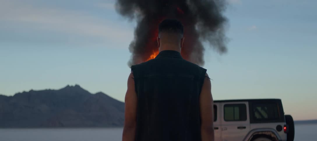 A man is standing outside watching a jeep burning in flames and there are mountains in the distance