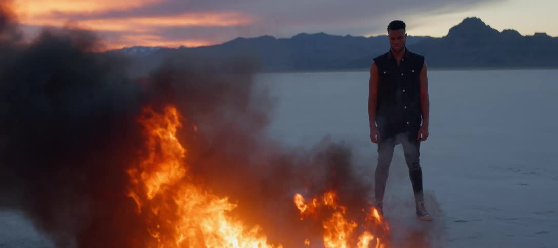 A man watches as a flame burns in the desert at sunset