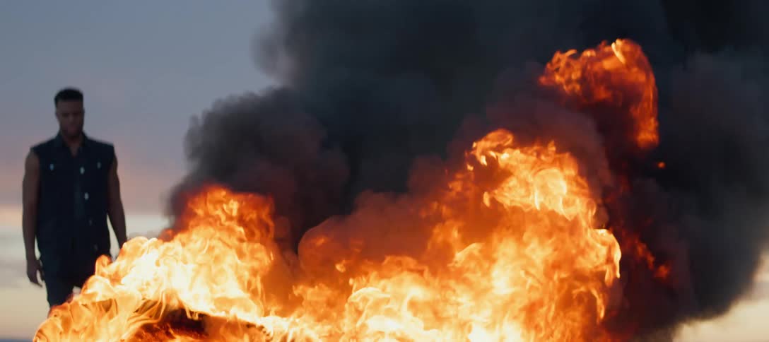 A man is standing outside watching and automobile burn in a raging fire as black smoke rises from the flames