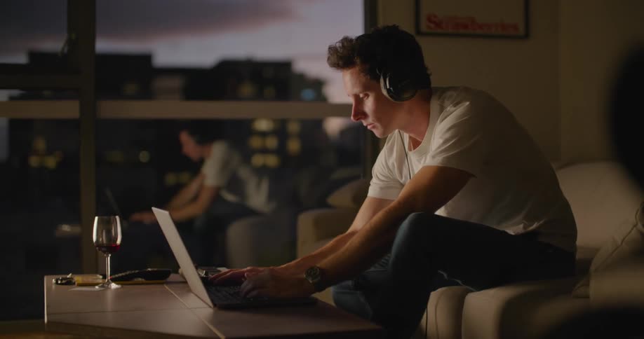Man sitting on the couch in the living room uses a laptop while wearing headphones at evening