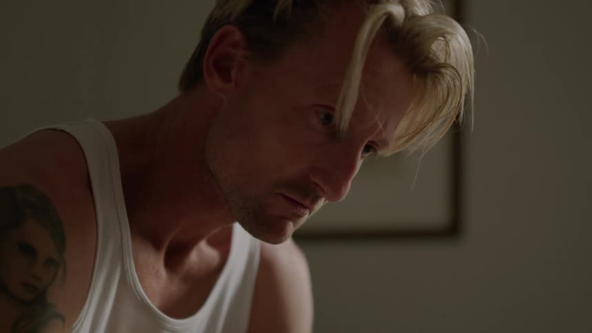 A man in an undershirt sits indoors while lost in thought