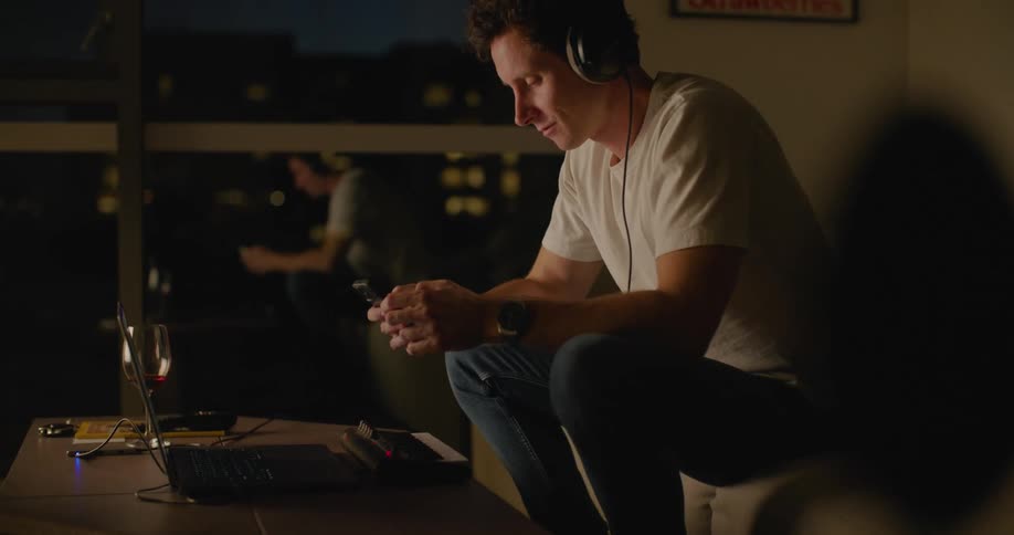 Man win headphones white t shirt and jeans sits on couch using smartphone in the living room at night