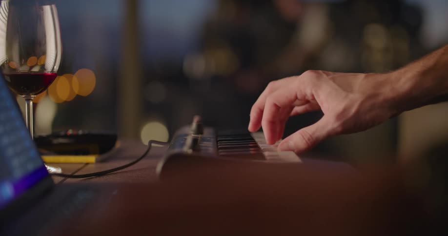 A man plays a keyboard with a laptop set up and a glass of wine