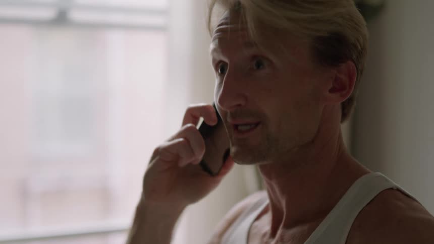 A man stands indoors next to a window while talking on his cell phone