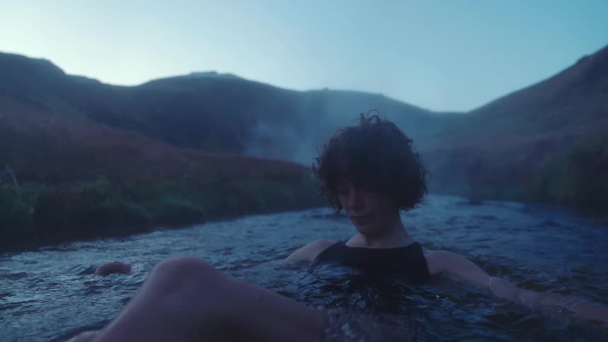 Woman with curly hair and black swimsuit sits in stream with mountains in the background