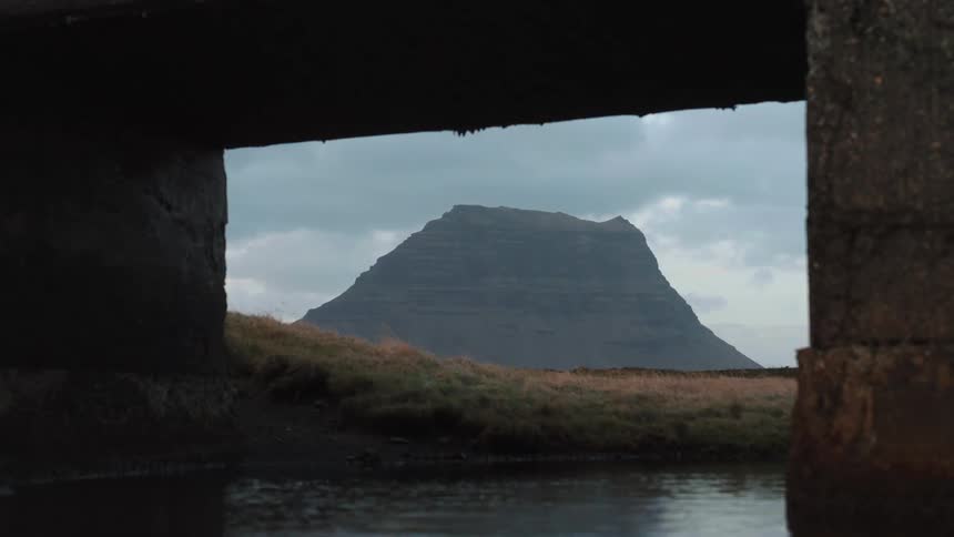 Iceland stream flows under an overpass and there is a mountain in the distance under a cloudy sky - Kirkjufellsfoss