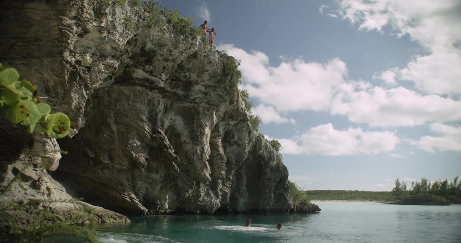 A pair of boys swim in the waters beneath a cliff as another pair looks down at them from the cliff top during the day