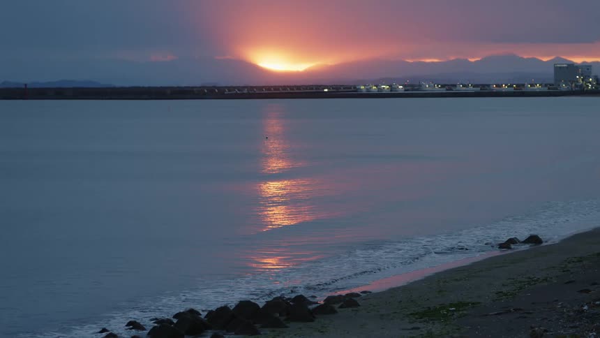 The sun sets behind some mountains while gentle waves lap the beach