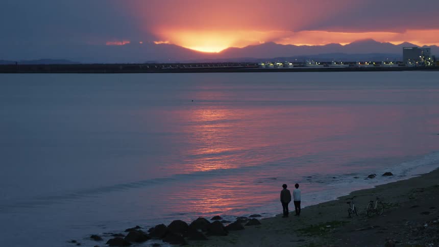 Two people standing on a sandy beach and watching the sun set