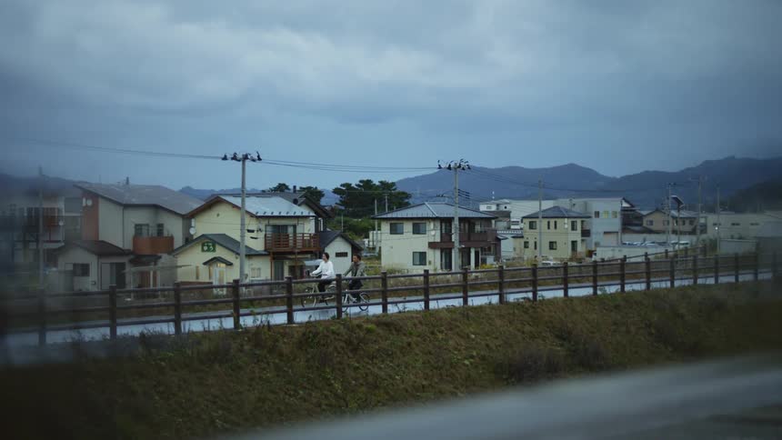 Two people riding bikes near buildings on a cloudy rainy day