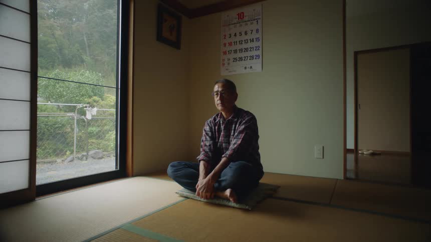 An older man is sitting down indoors and is doing yoga