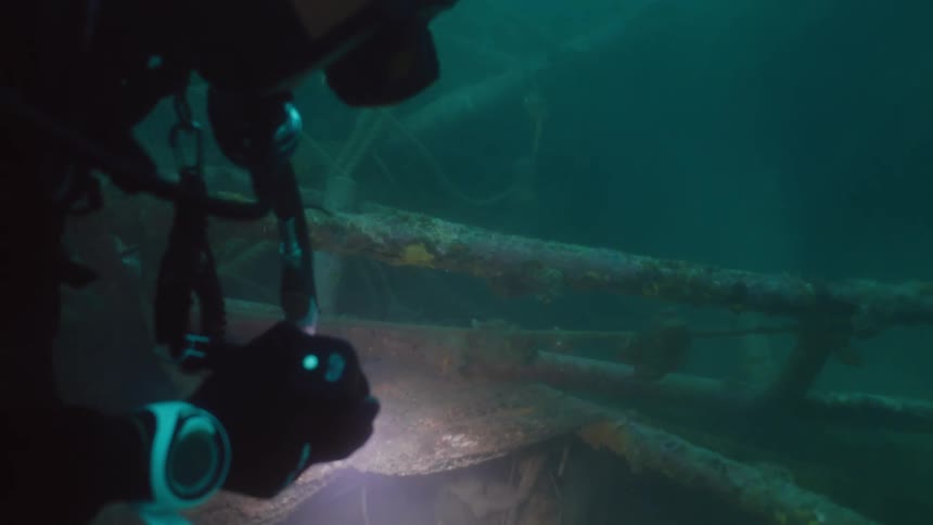 Scuba diver with flashlight examines wreck on seafloor in murky water
