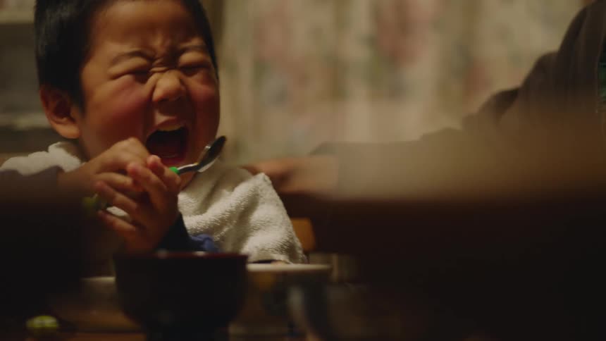 Young asian boy in white bib eats his meal with a spoon with adult by his side