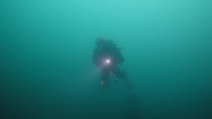 A scuba diver swimming in murky water with a flashlight