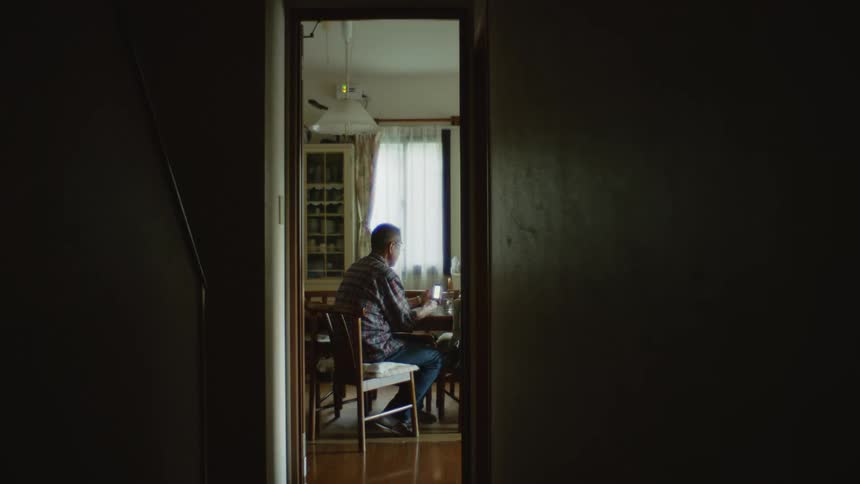 An older white man is sitting down inside of his home