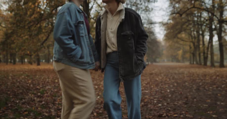 Two women are holding hands as they walk through a park with trees in fall colors and leaves covering the ground
