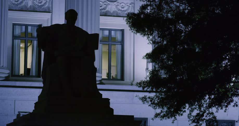 Sculpture of sitting person is silhouetted against white government building with classical architecture at night