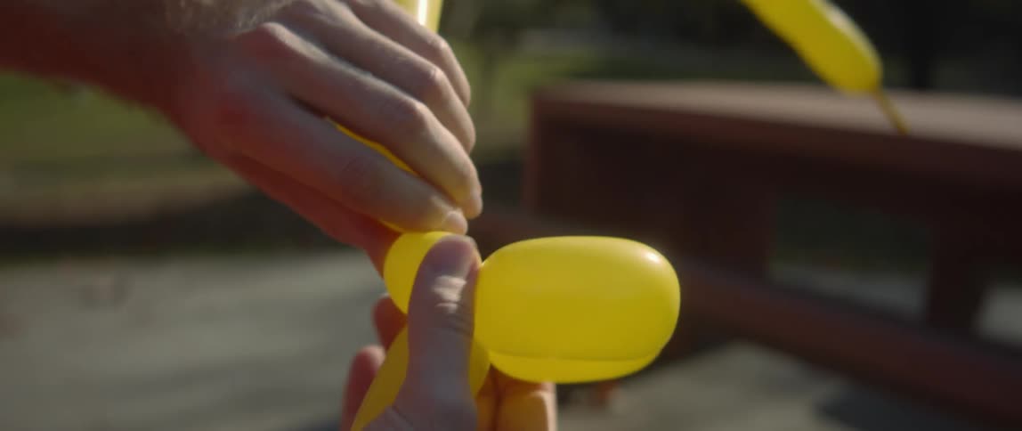 The sun shines on a man outside twisting a yellow balloon into an animal with his hands