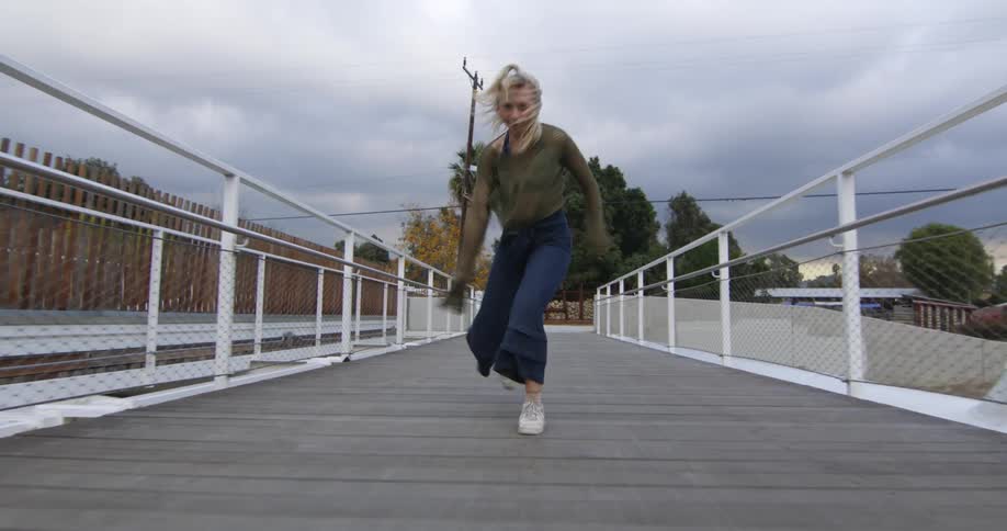 Blonde dancer dances along wooden pedestrian bridge under an overcast sky near a transmission tower