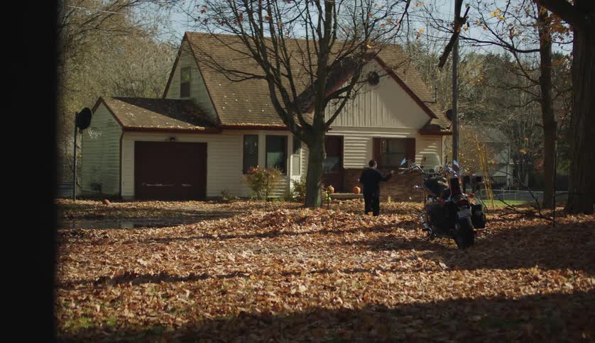 A young man raises his arms to keep his balance while walking over a large mound of fallen leaves to reach a house during the day