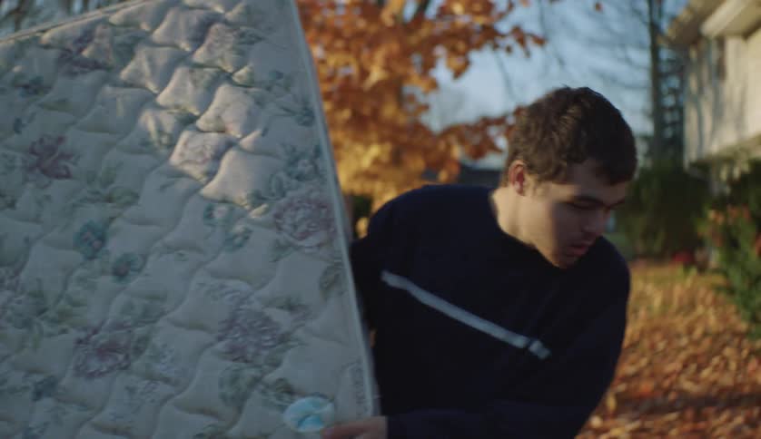 Man in dark blue sweater shakes his hands and removes mattress in the yard with brown leaves on the ground