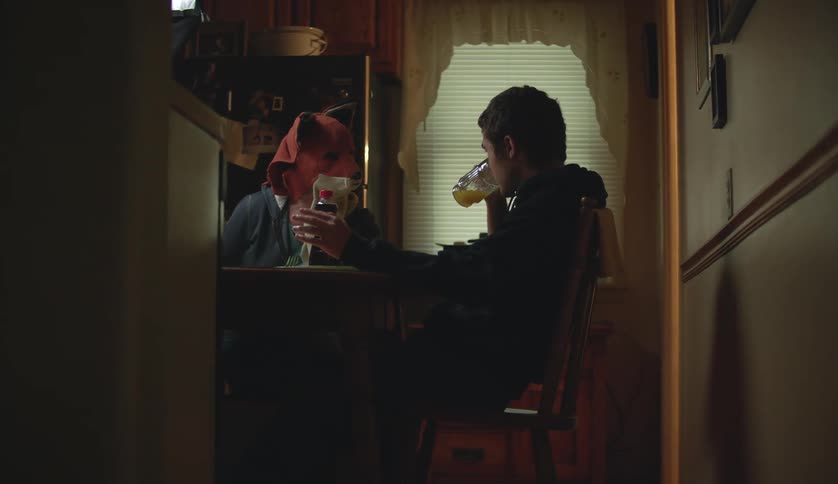 A teenage boy is sitting at the kitchen table drinking a glass of orange juice when someone wearing an animal head mask walks in and sits at the table