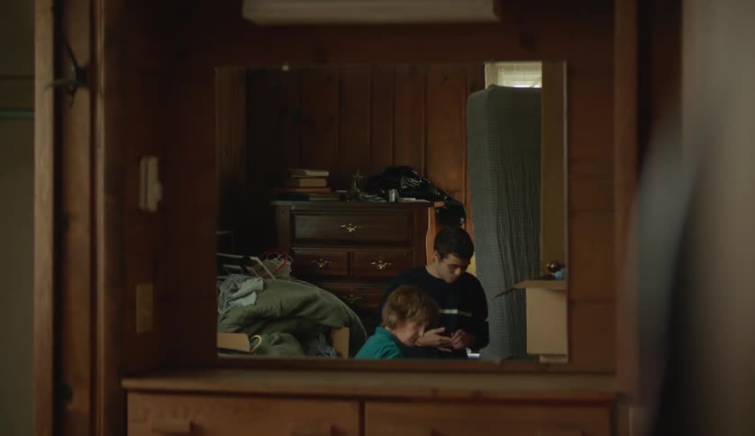 The reflection of a teenage boy and his mother are on the floor of a room sorting things out are seen in a mirror