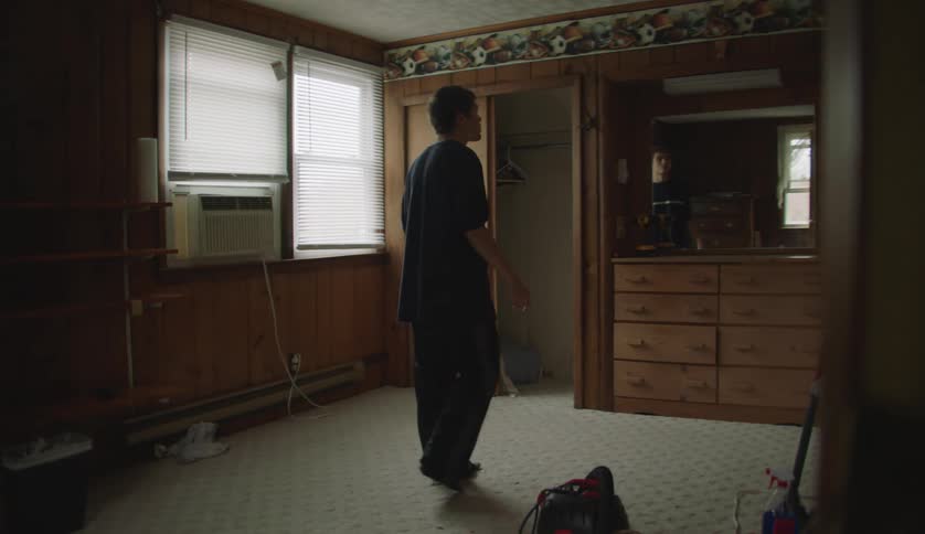 A teenage boy walks into a room and looks around as his reflection is in the dresser mirror