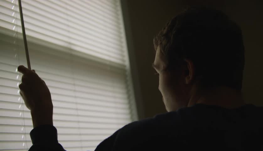 A young white man is inside of his house and looking out the window