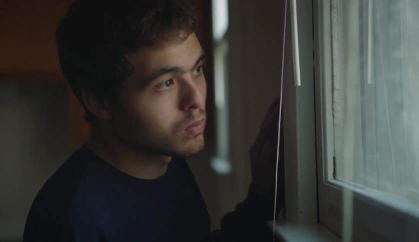 A young man hiding near a window tilts his head to peek outside