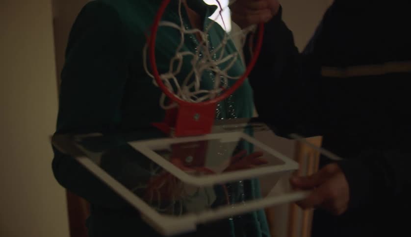 A teenage boy is standing in a room and his mother hands him a basketball hoop