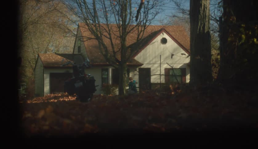 A person wearing a turquoise sweater is sweeping the front porch of a house during a sunny autumn day