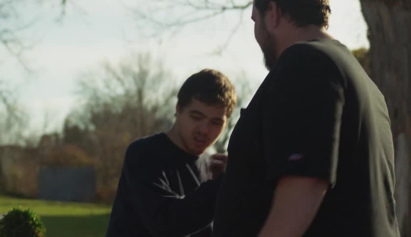 Two men shake hands near table at yard sale while person swings in background