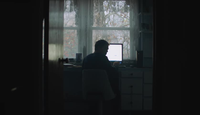 A man is sitting in front of a computer on a desk near windows as he turns around and looks away from the screen