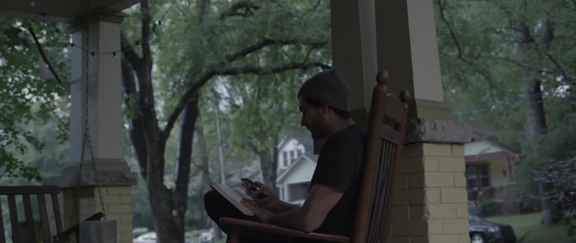 A man sits on his porch reading a book and phone