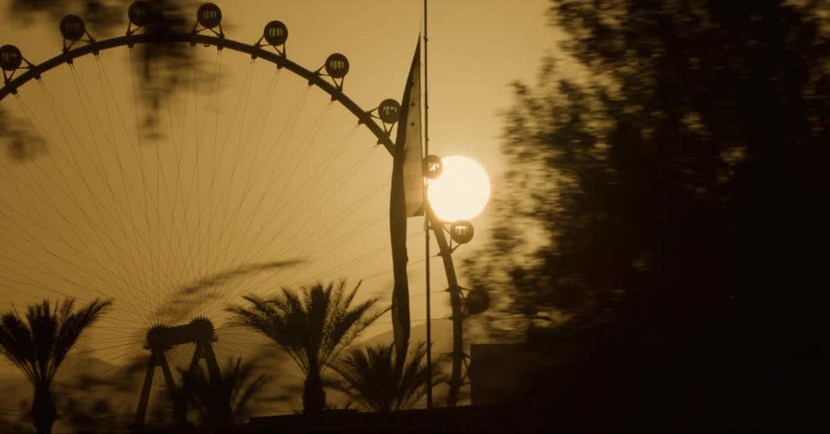 Brush ferris wheel and buildings are silhouetted against red sky as sun sits above mountains