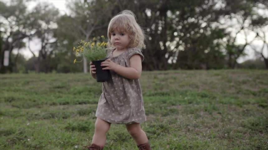 A little girl walking through a field as she carries a pot of yellow flowers